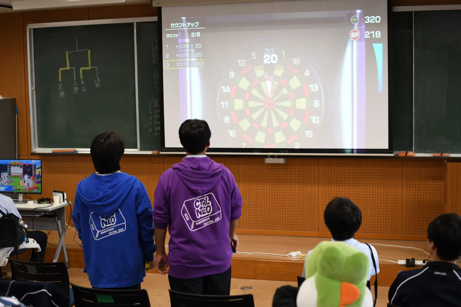 スクリーンに映したダーツのゲームを、お揃いのパーカーを着た学生が見ている様子